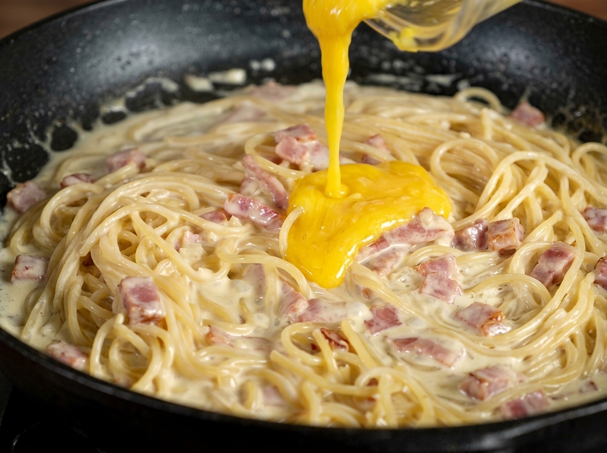 Pâte jaune de jaune dœuf et parmesan versée sur les spaghetti à la crème dans une poêle sombre.