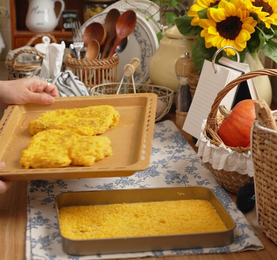 Mains tenant une plaque de cuisson avec deux grandes escalopes de porc panées dans une cuisine chaleureuse.
