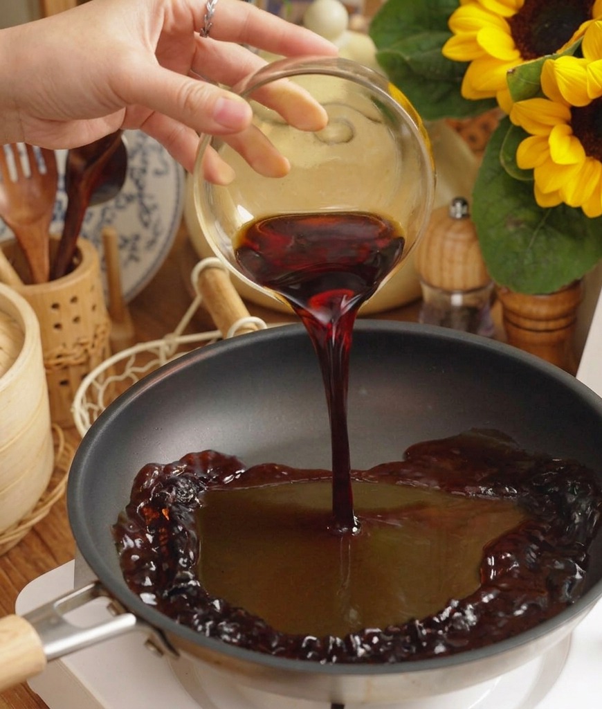 Une sauce foncée savoureuse versée dun bol en verre dans une poêle noire sur une plaque électrique blanche.