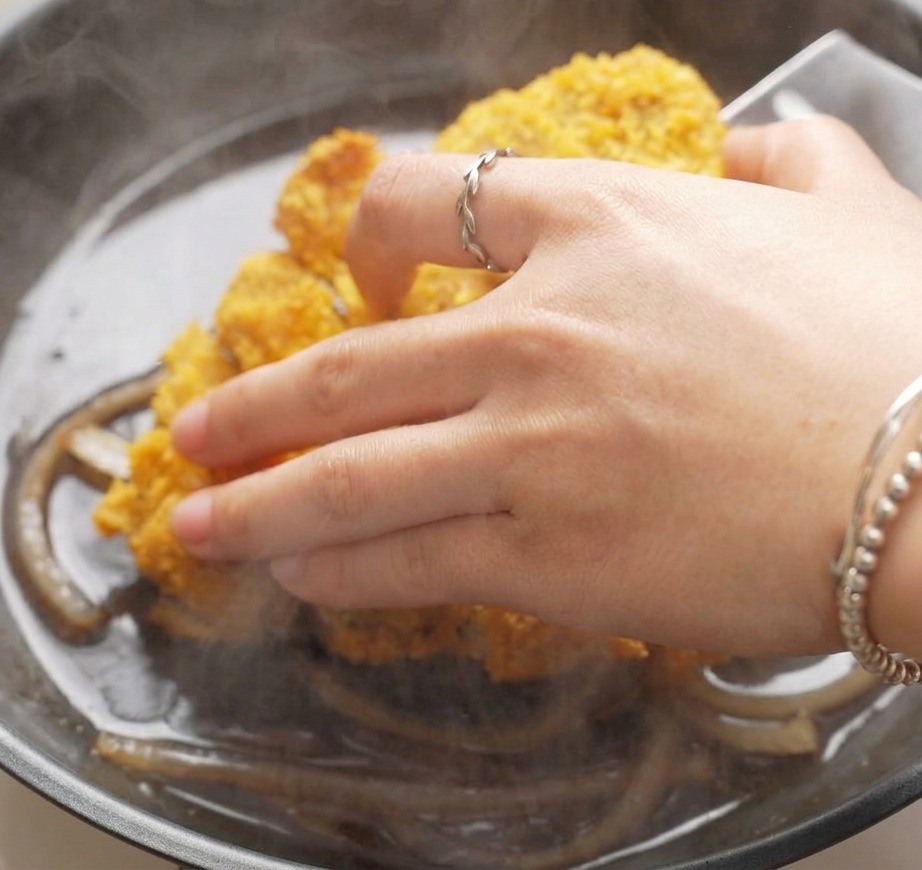 Une main déposant des tranches descalope de porc croustillante dans une poêle remplie doignons mijotés et dune sauce foncée.