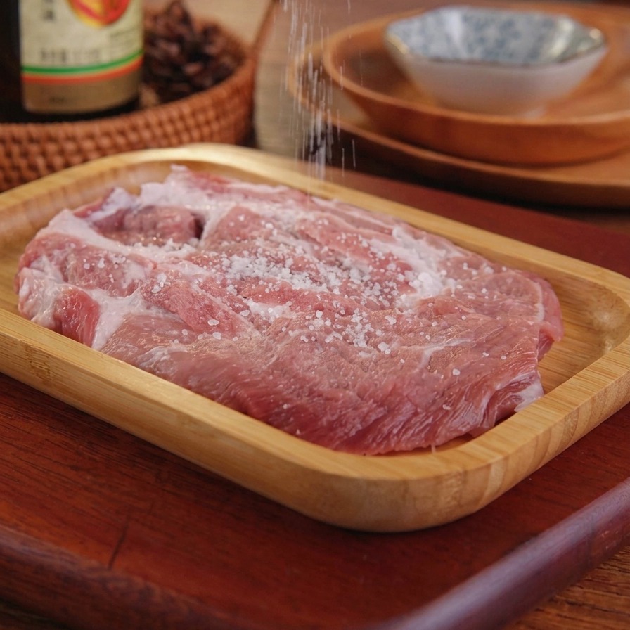 Du sel saupoudré à la main sur une côtelette de porc crue placée sur un plateau de service en bois.