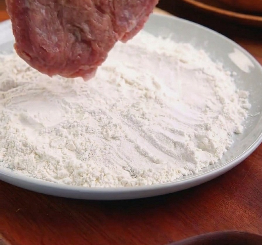 Une côtelette de porc tenue par des pinces métalliques que lon plonge dans une assiette de farine blanche.