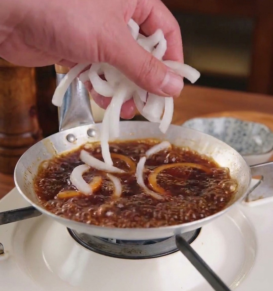 Une main ajoutant des lamelles doignon blanc frais dans une sauce brun foncé frémissante dans une poêle métallique peu profonde.