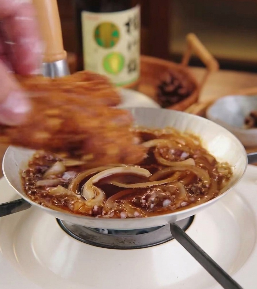 Une côtelette de porc panée et dorée déposée sur un lit doignons mijotant dans leur sauce au sein dune petite poêle.