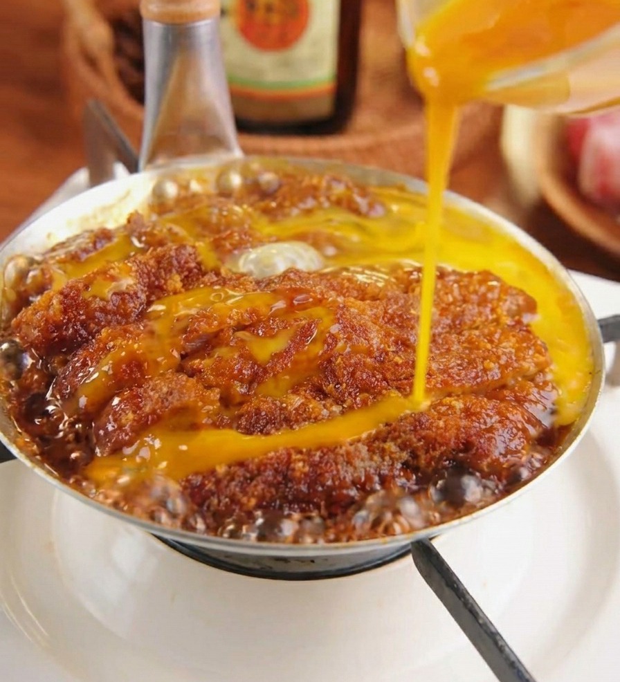 Des œufs battus versés sur une côtelette de porc panée qui mijote dans une sauce sombre avec des oignons.