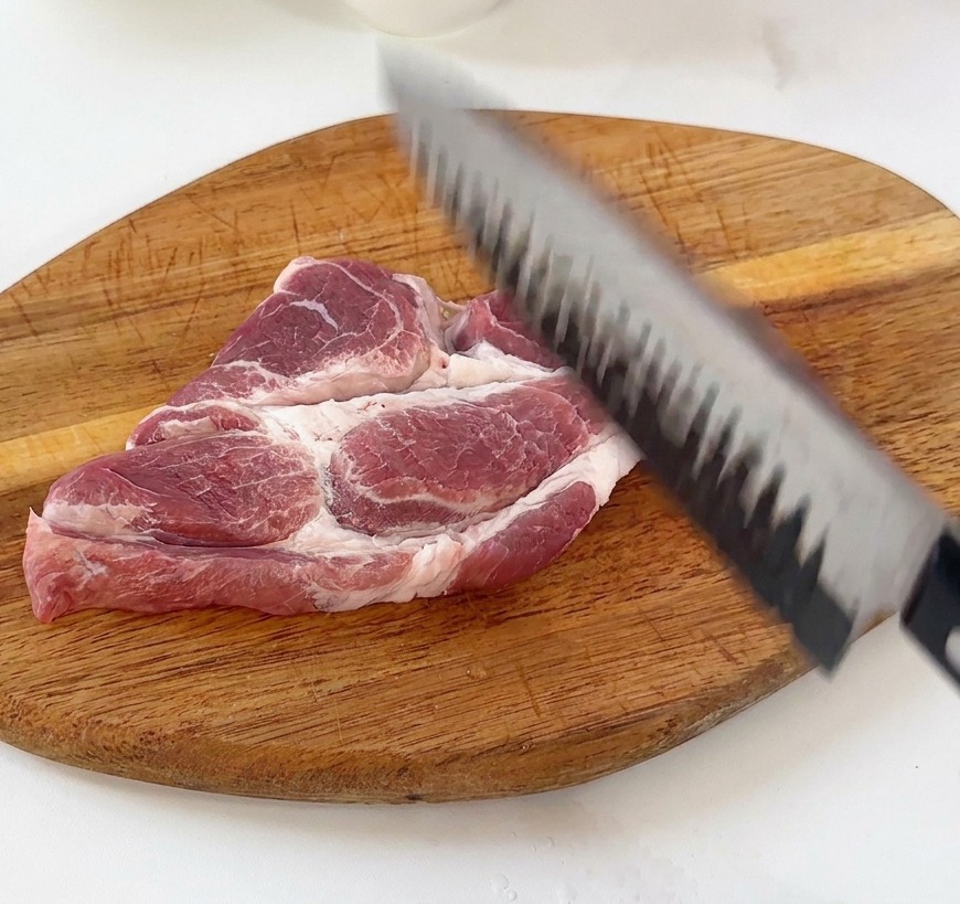 Une main utilisant le dos dun couteau de chef pour attendrir une côtelette de porc crue sur une planche en bois.