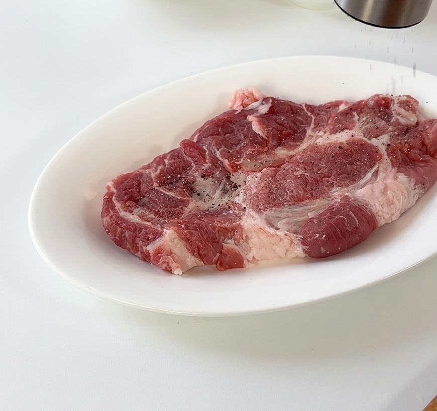 Poivre noir moulu sur une côtelette de porc crue reposant sur une assiette en céramique blanche.