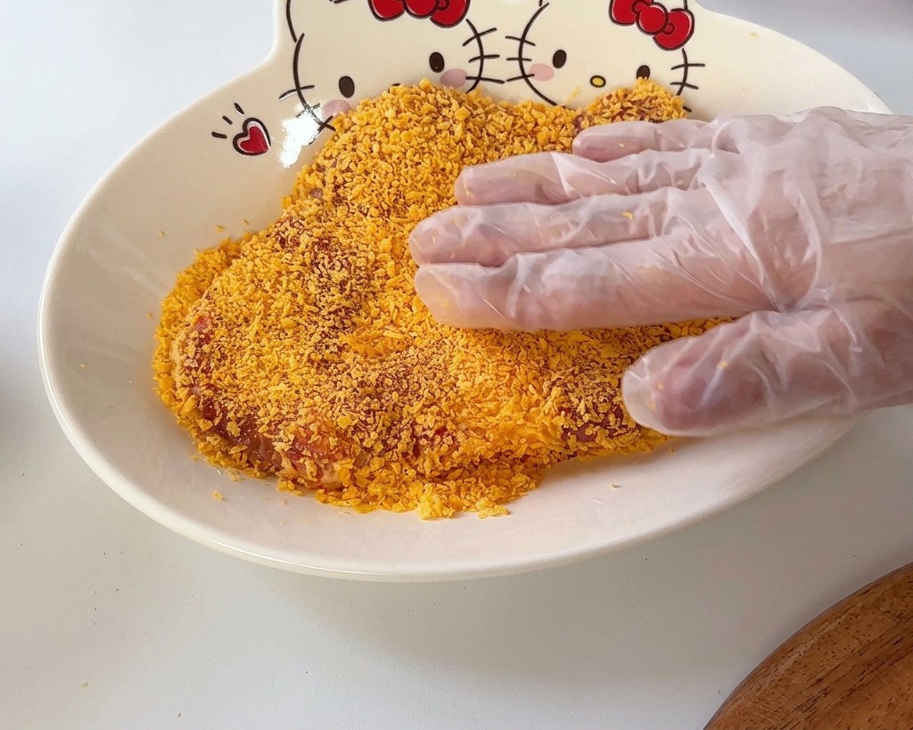 Une main pressant une côtelette de porc dans une couche de chapelure orangée à lintérieur dun bol motif Hello Kitty.