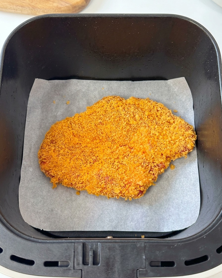 Une côtelette de porc panée posée sur du papier sulfurisé dans le panier noir dune friteuse sans huile.