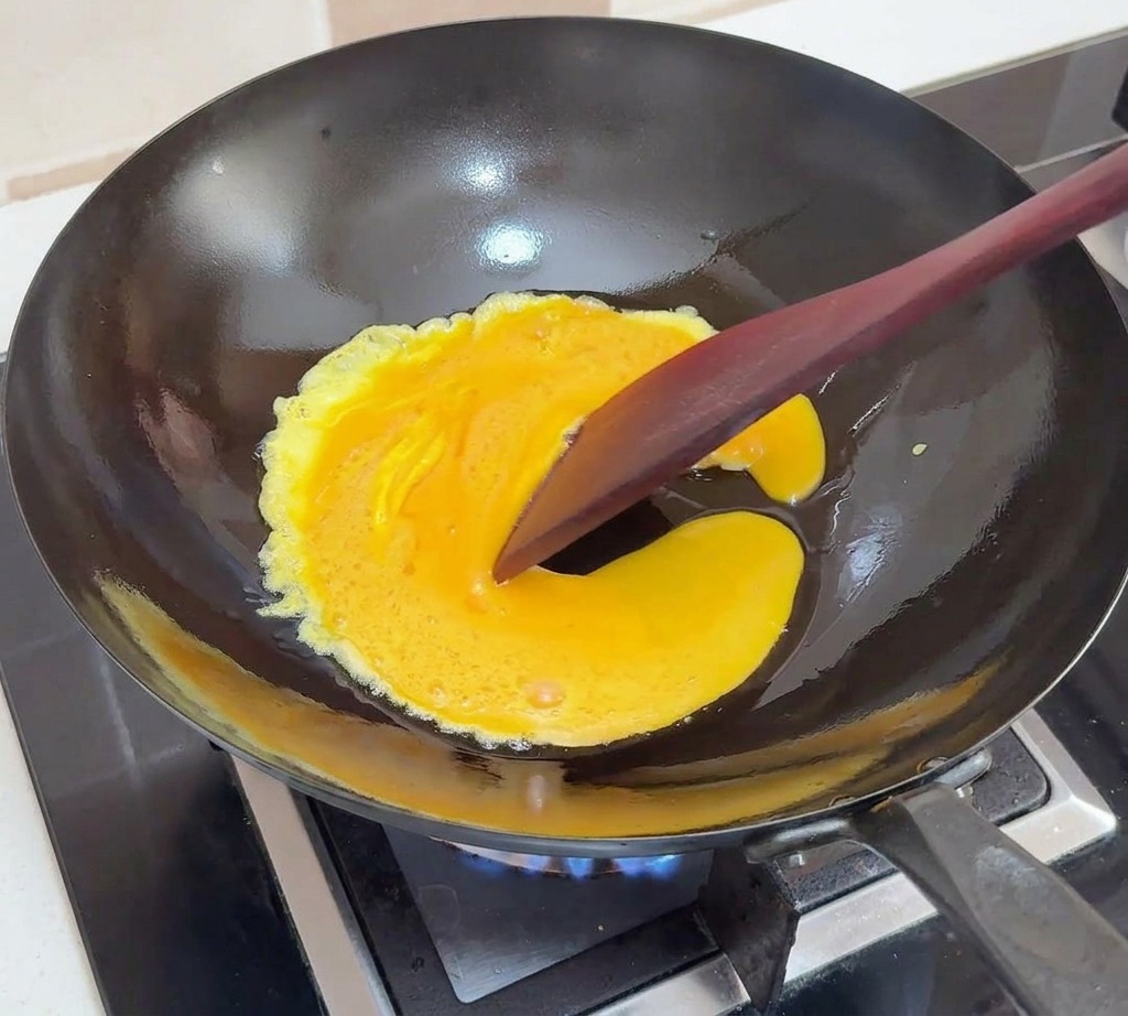 Des œufs battus cuisant dans un wok noir, ramenés vers le centre avec une spatule foncée pour créer des plis souples.