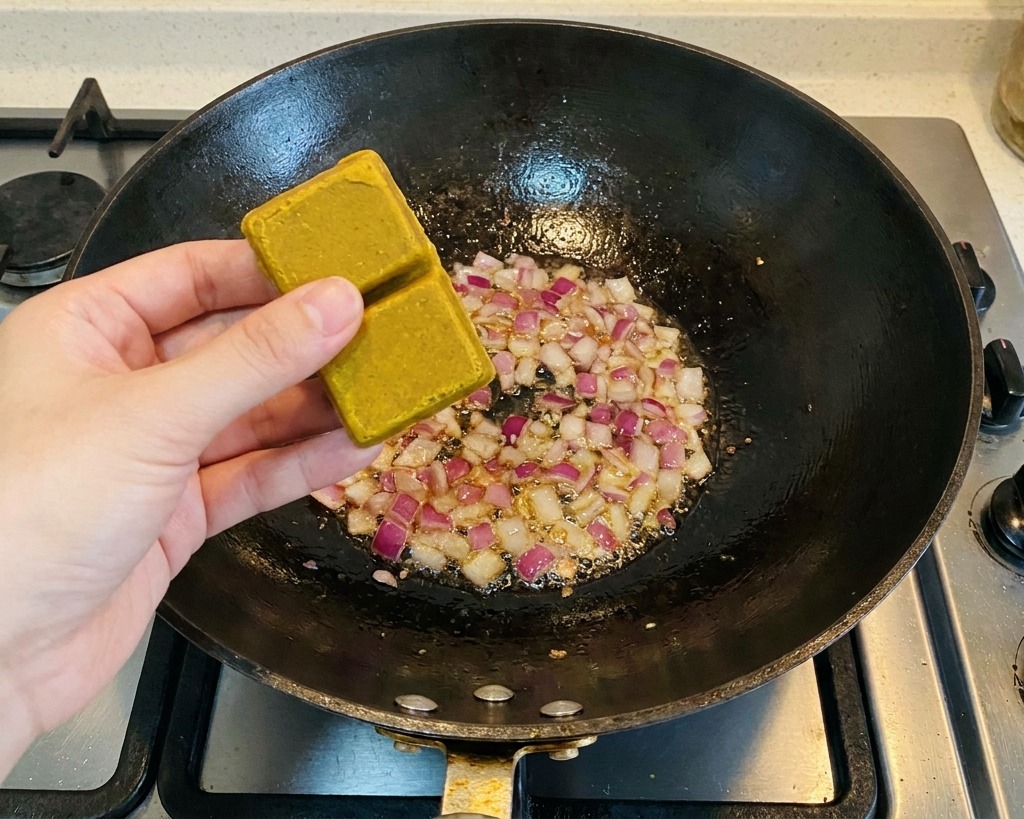 Une personne tenant deux blocs de curry brun au-dessus dun wok contenant des morceaux doignon rouge sautés.