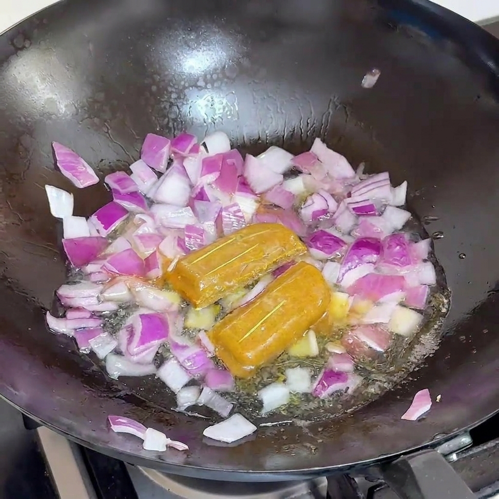 De leau versée dans un wok noir rempli doignons rouges et de deux blocs entiers de curry.