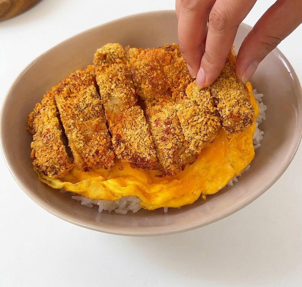 Des tranches de porc pané déposées sur une omelette jaune baveuse dans un bol en céramique beige rempli de riz blanc.