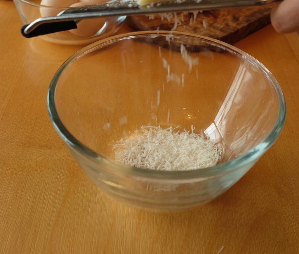 Un bloc de Pecorino Romano râpé finement dans un bol en verre, créant une montagne de copeaux blancs.