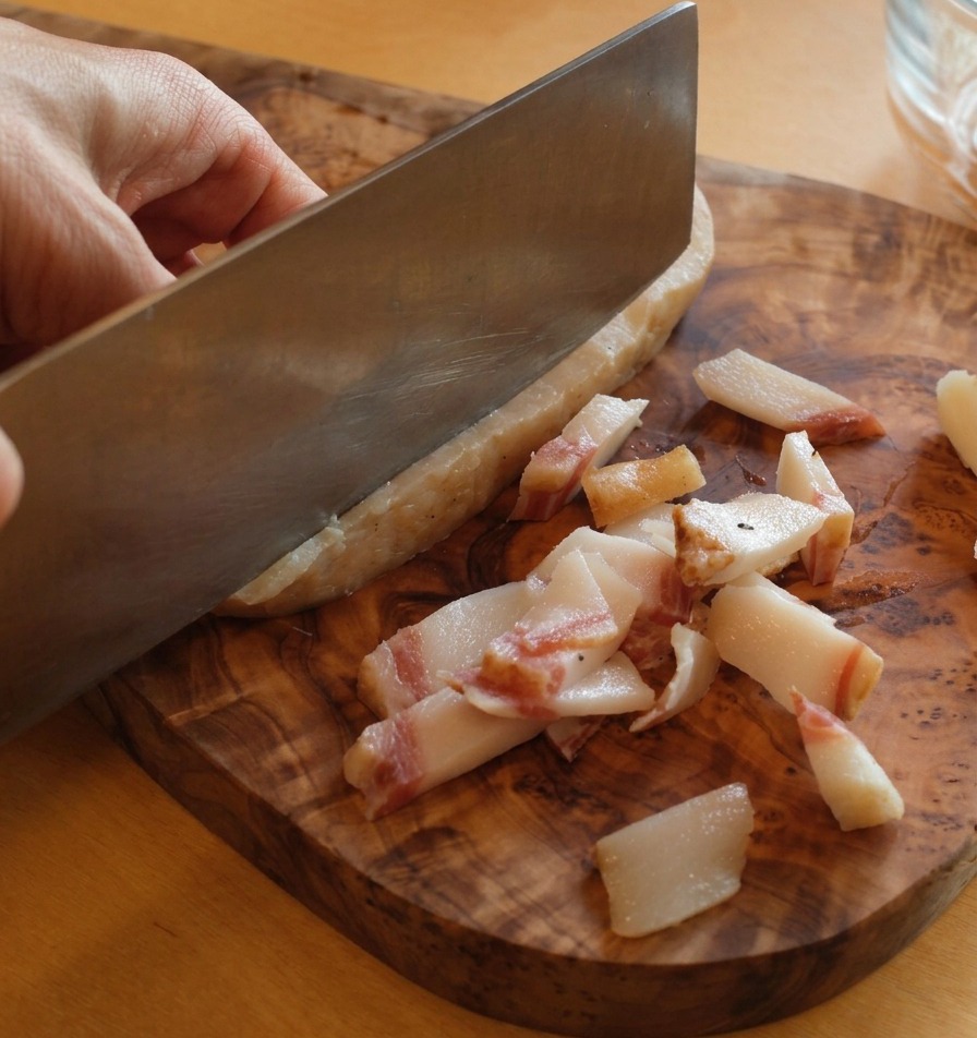 Un couteau aiguisé retirant la peau dure et sombre dun morceau de guanciale sur une planche en bois.