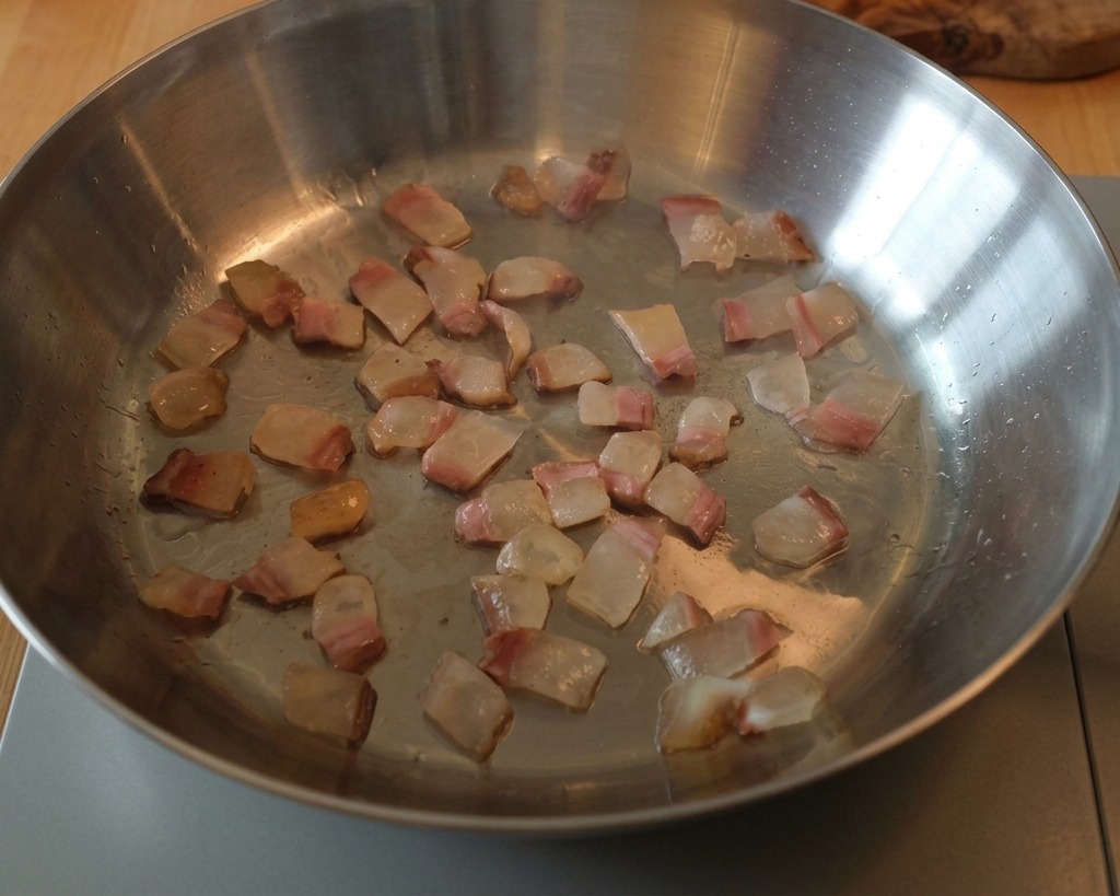 Dés de guanciale cuisant dans une poêle en inox, le gras se transformant en liquide translucide.
