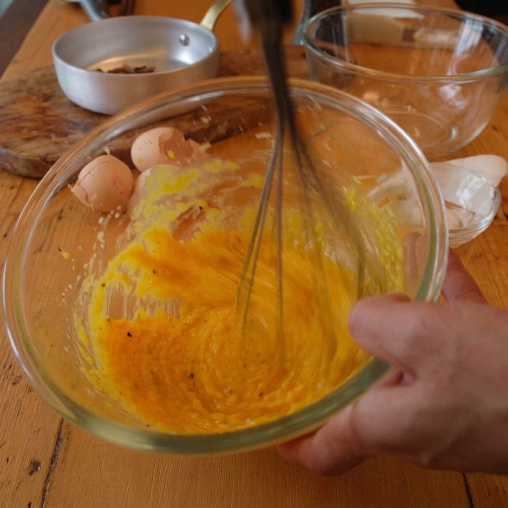 Une main mélangeant la sauce aux œufs pendant que de leau de cuisson chaude est ajoutée progressivement.