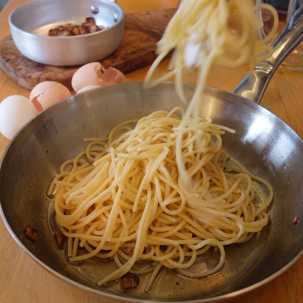 Une pince déposant des spaghettis cuits dans une poêle en inox contenant le gras du guanciale.