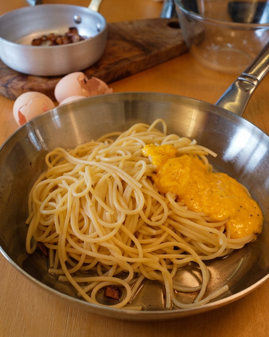 Une sauce dorée aux œufs et au fromage versée sur un dôme de spaghettis fumants dans une poêle.