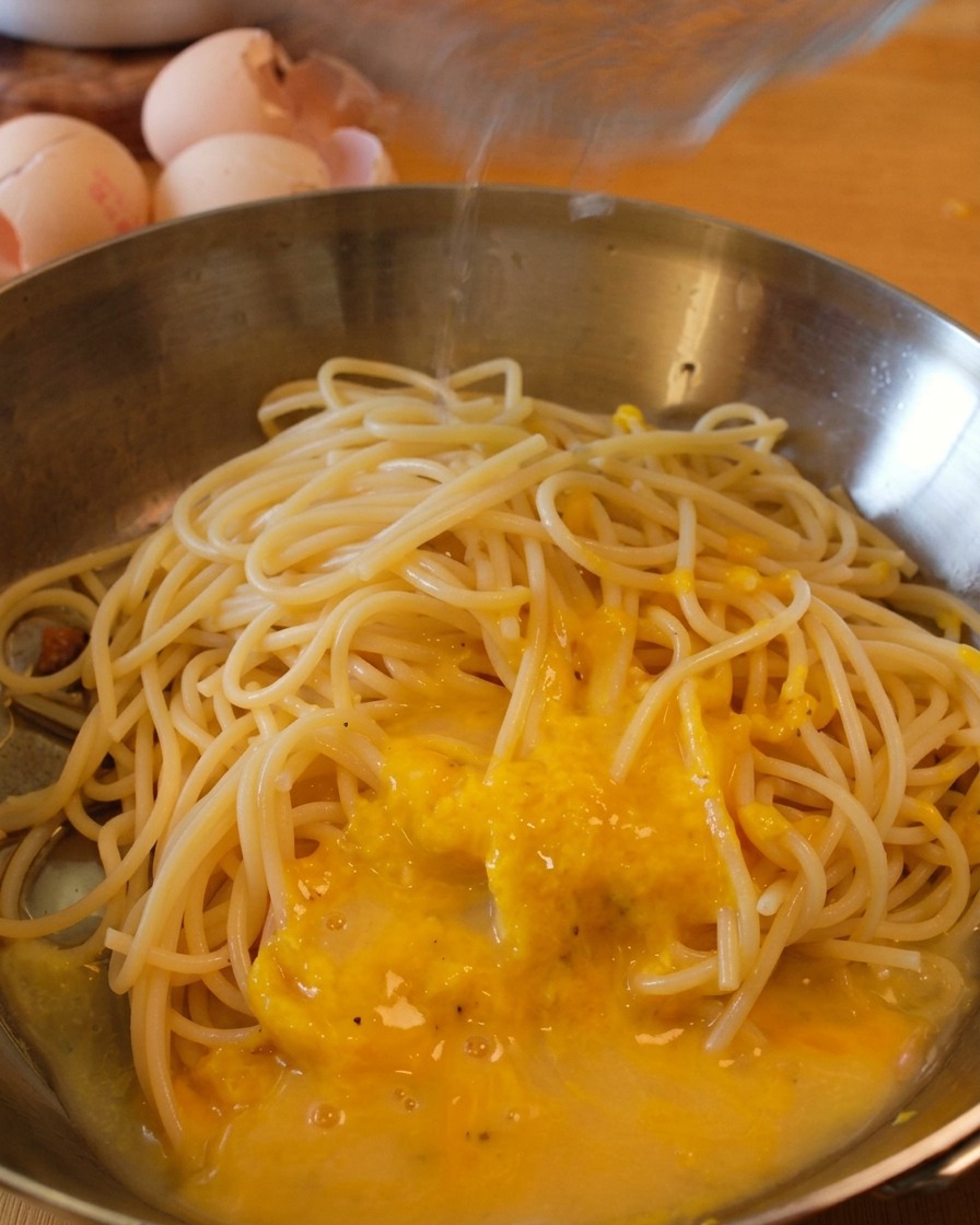 Spaghettis dans une poêle avec la sauce aux œufs alors quun filet deau de cuisson est ajouté.