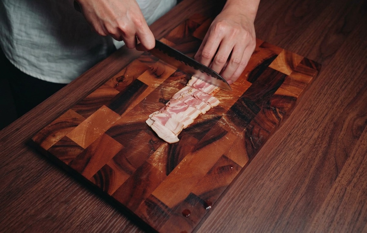 Une personne utilisant un couteau de chef pour couper des tranches de lard marbré en petits rectangles sur une planche en bois.