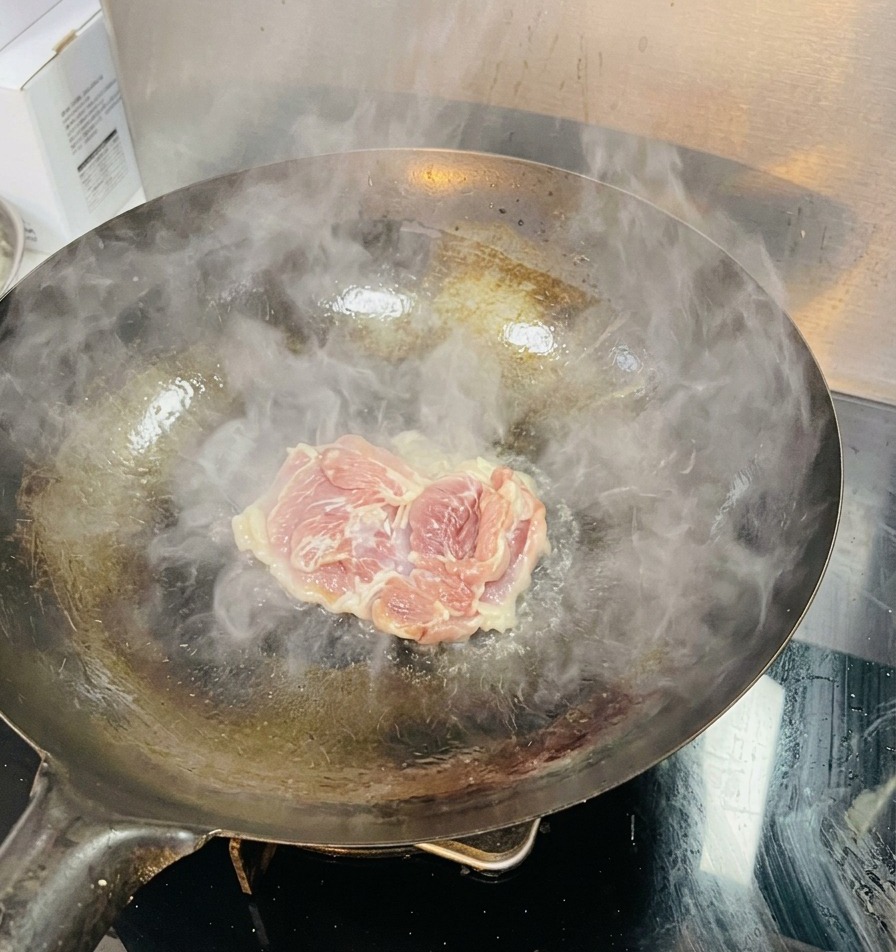 Une cuisse de poulet crue déposée dans un wok très chaud, dégageant de la vapeur dans une huile frémissante.