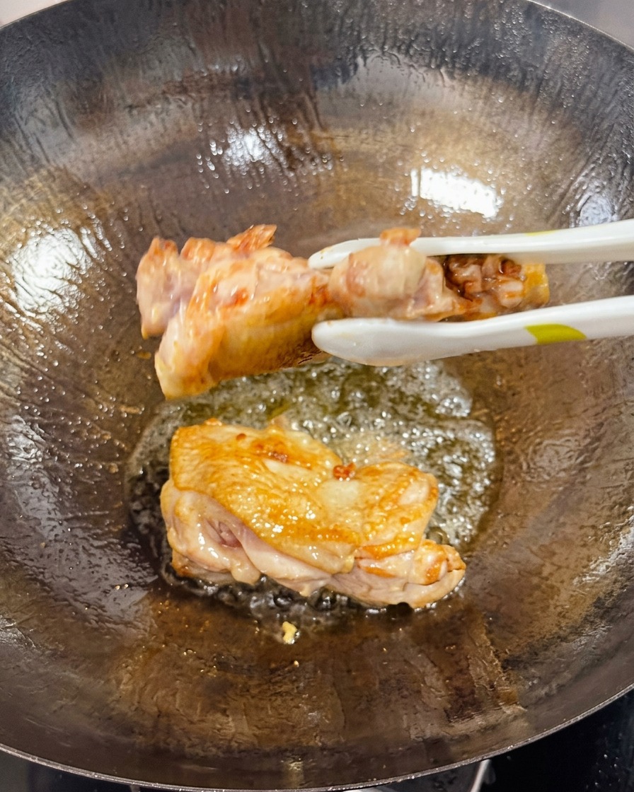 Des pinces retournent une cuisse de poulet saisie dans un wok, montrant le côté cuit bien doré et croustillant.
