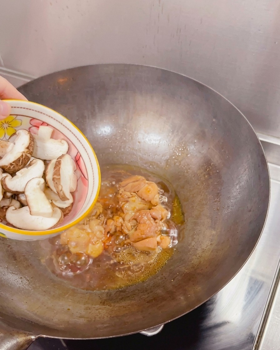 Des champignons shiitaké frais et émincés ajoutés depuis un bol dans un wok contenant le poulet saisi et la sauce frémissante.