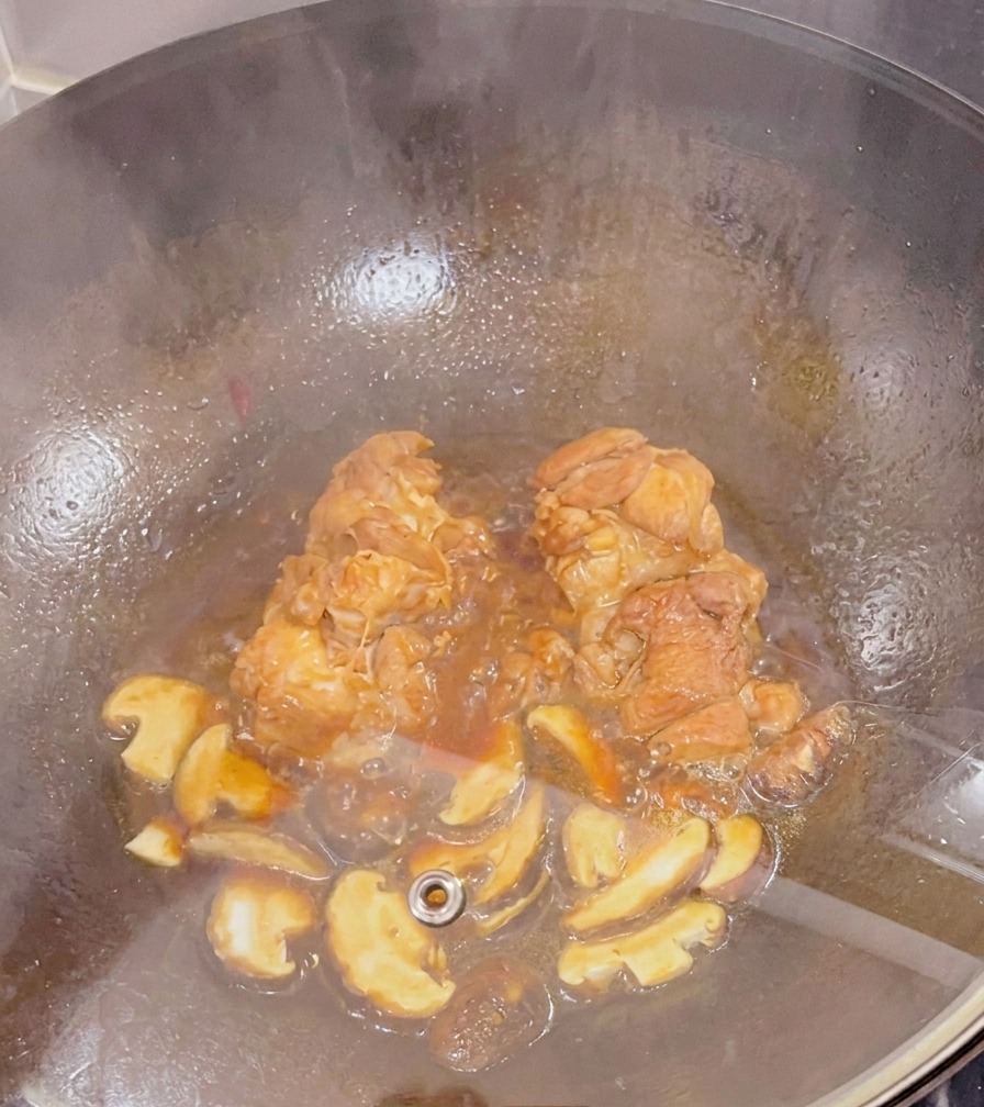 Des cuisses de poulet et des champignons shiitaké émincés mijotant dans une sauce sombre à lintérieur dun wok couvert par un couvercle en verre.