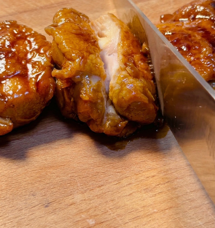 Un couteau aiguisé tranchant une cuisse de poulet teriyaki très caramélisée sur une planche en bois, révélant une viande juteuse à lintérieur.