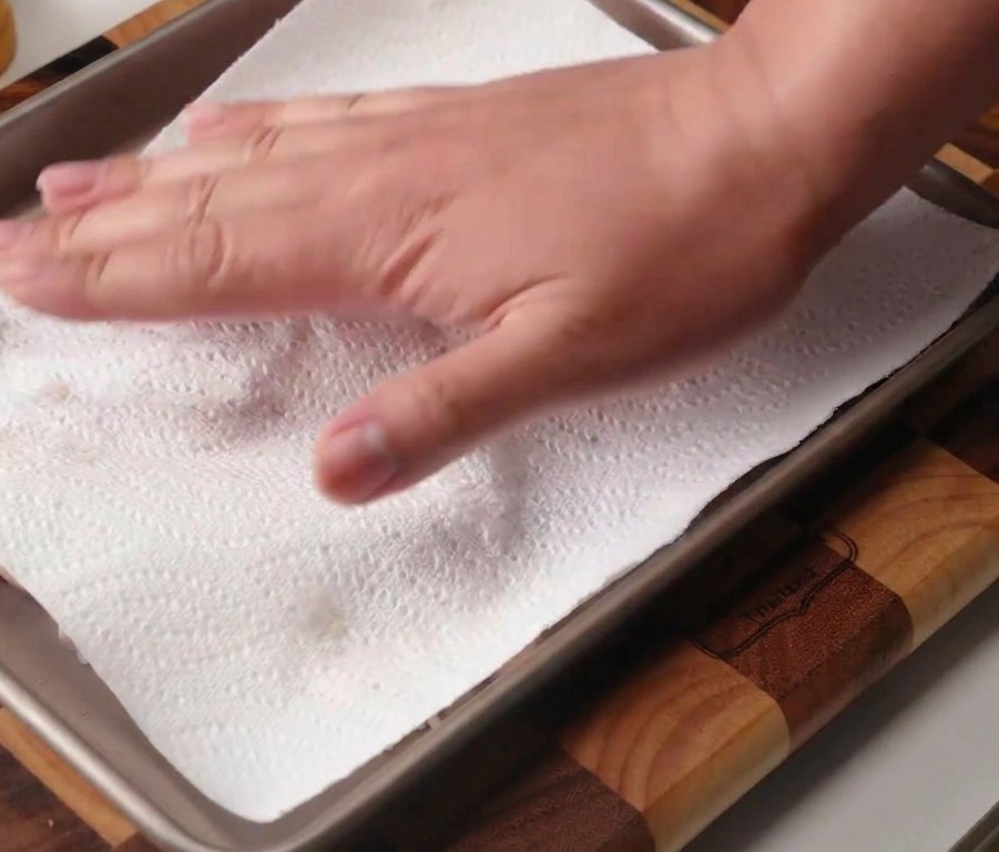Une main utilisant du papier absorbant blanc pour sécher des hauts de cuisse de poulet crus disposés sur une plaque de cuisson en métal.
