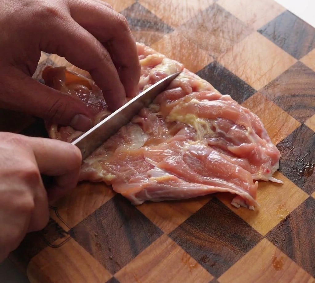 Une main utilisant le dos dun couteau de chef pour aplatir des hauts de cuisse de poulet désossés sur une planche à découper en bois quadrillée.