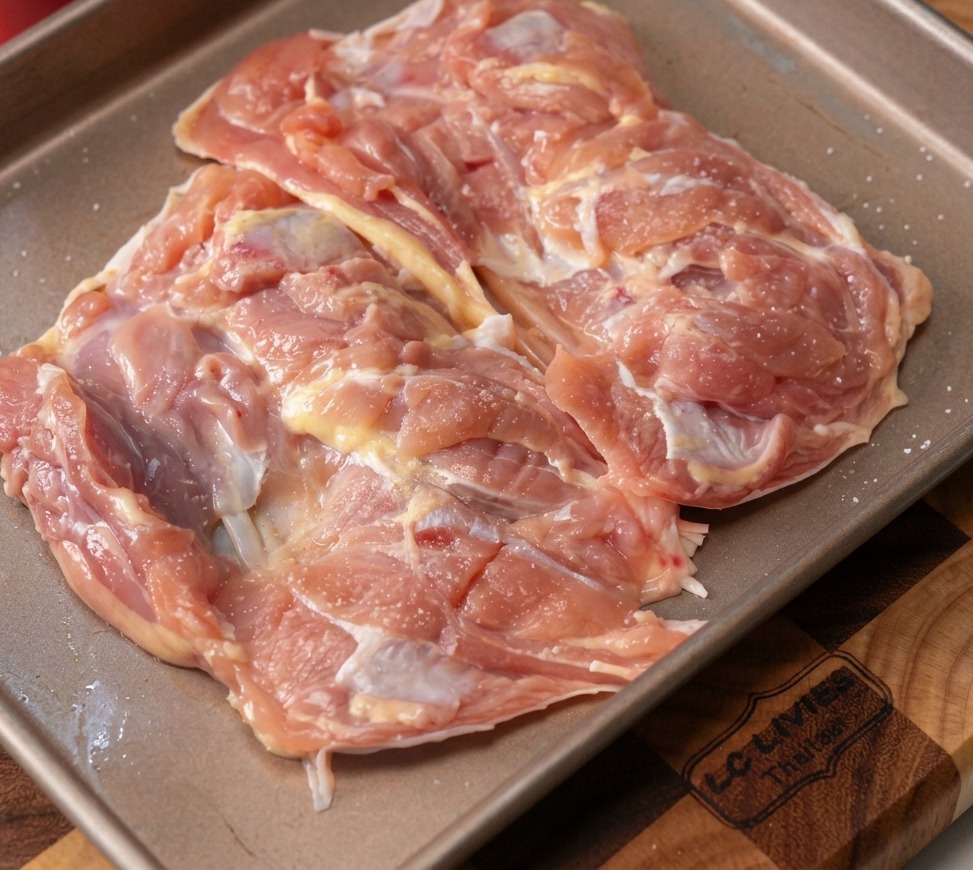 Hauts de cuisse de poulet crus désossés sur une plaque de cuisson en métal, parsemés dail émincé et de poivre noir.