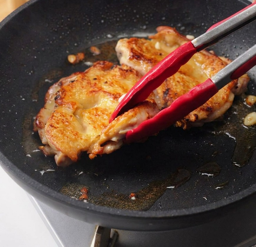 Une main versant une sauce teriyaki sombre depuis un bol en verre sur les hauts de cuisse de poulet saisis dans une poêle.