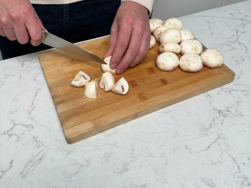 Mains utilisant un couteau de chef pour couper des champignons de Paris en quartiers sur une planche en bois.