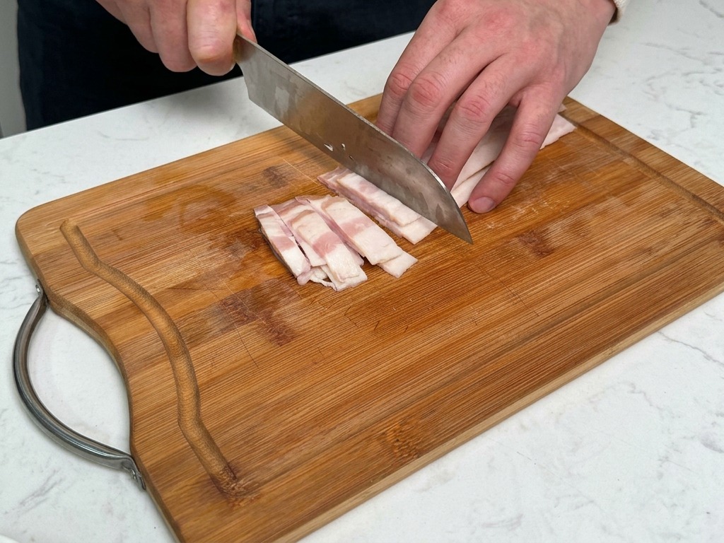 Une personne coupant dépaisses tranches de bacon en lanières sur une planche à découper en bois.