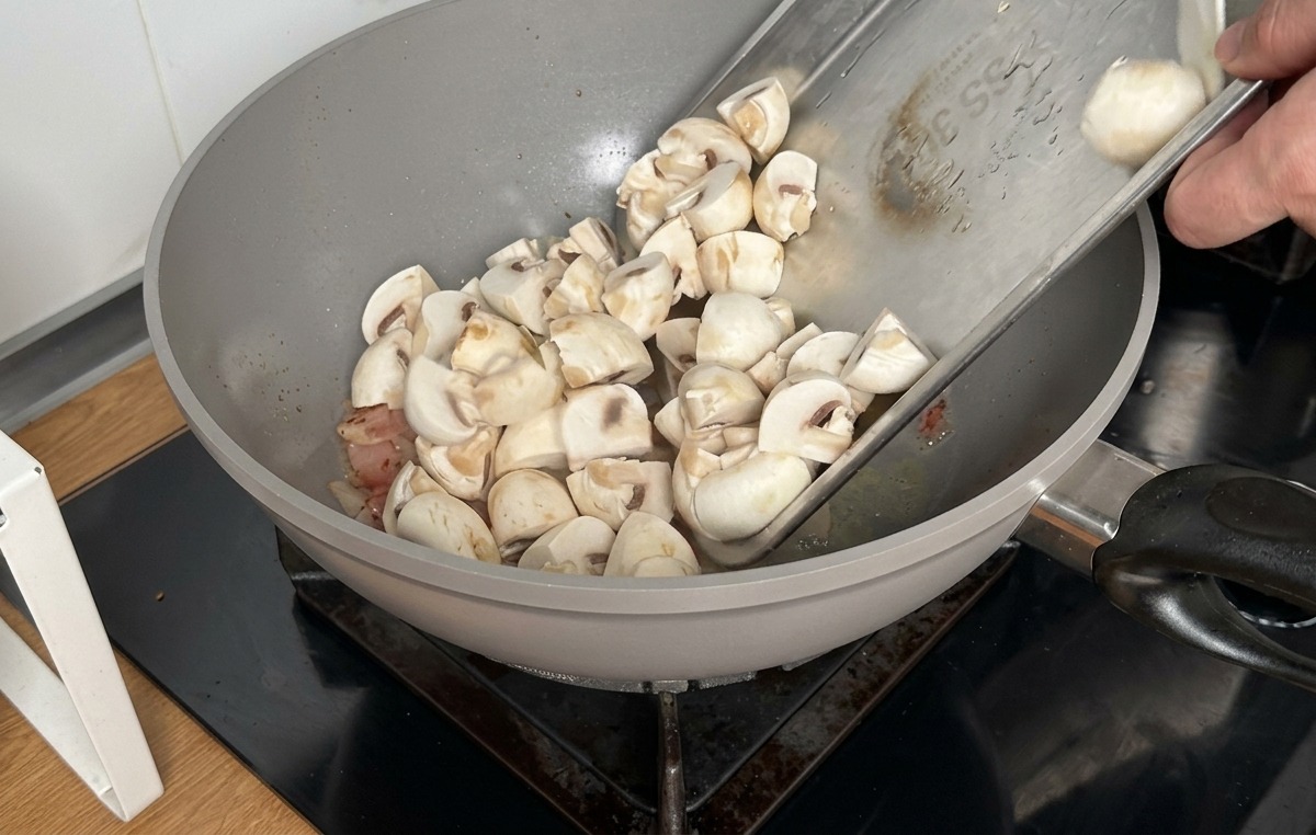Verser les quartiers de champignons de Paris depuis un plateau métallique dans un wok contenant le bacon cuit.