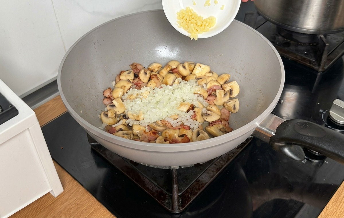 Verser des oignons et de lail hachés depuis un bol blanc sur les champignons et le bacon cuits dans un wok.