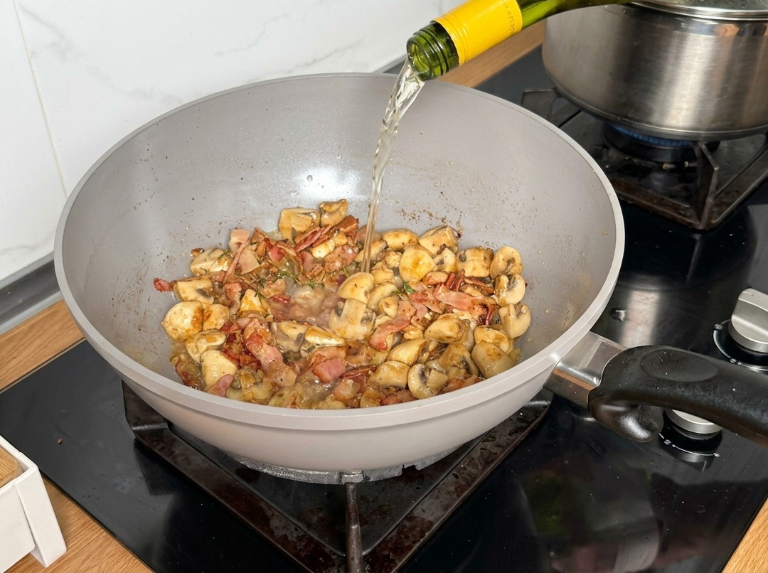 Vin blanc versé depuis une bouteille dans une poêle contenant les champignons et le bacon.
