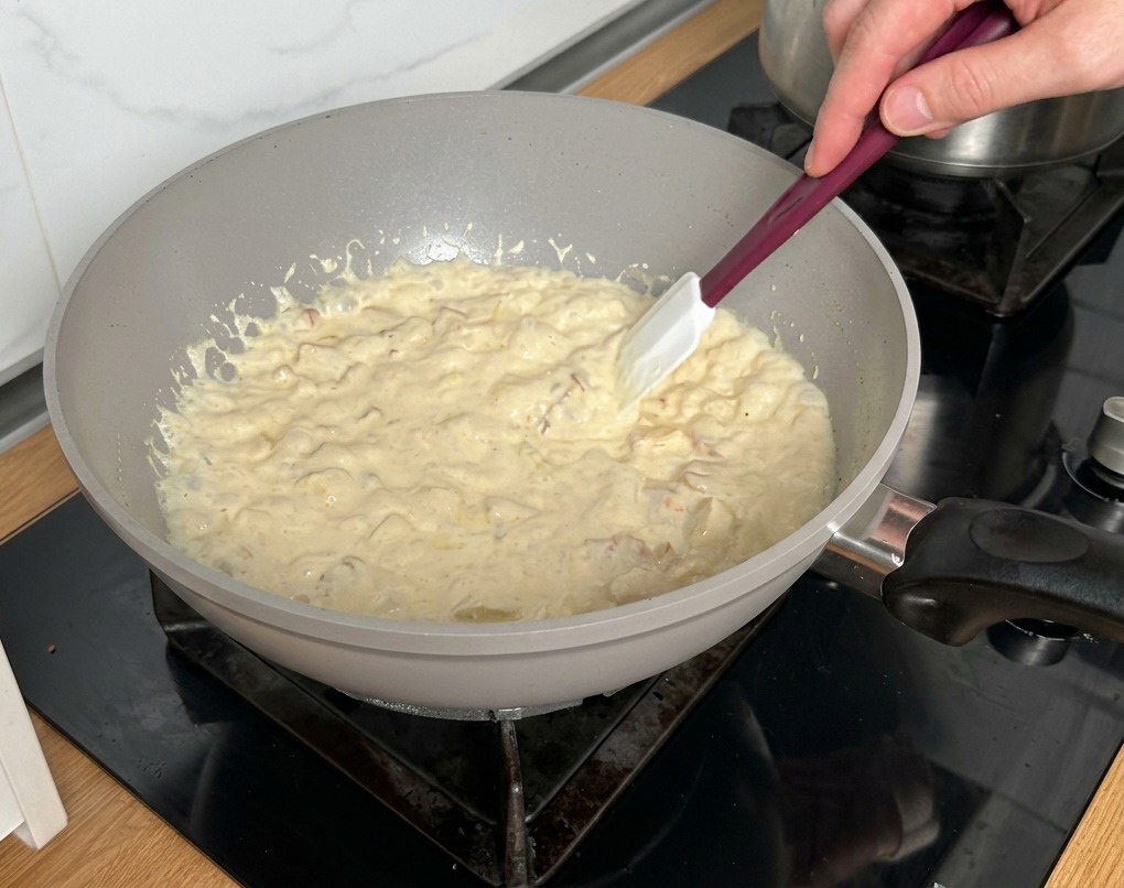 Une spatule blanche remuant une sauce crémeuse épaisse avec bacon et champignons dans une poêle claire.