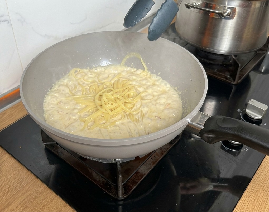 Pinces métalliques soulevant des pâtes cuites vers une poêle remplie dune sauce crémeuse au bacon et champignons.