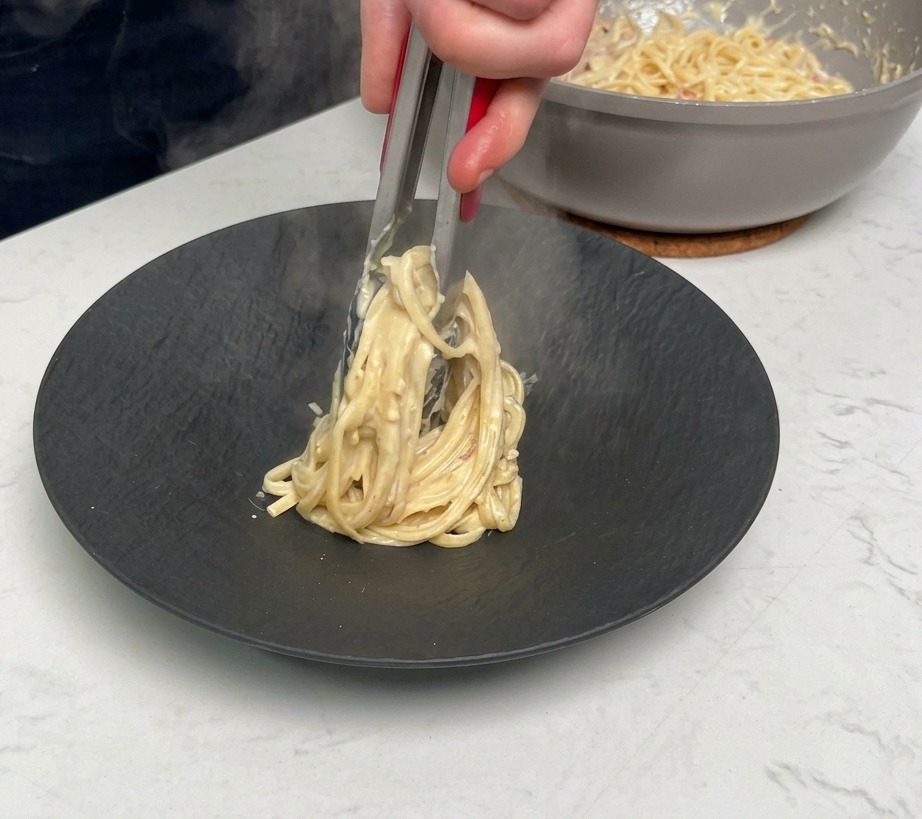 Utilisation de pinces métalliques pour enrouler et dresser une portion de pâtes crémeuses sur une assiette noire mate.