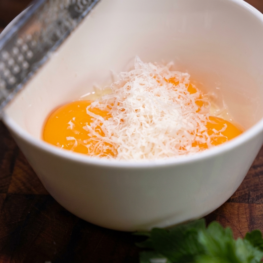 Une râpe en métal produisant de fins copeaux blancs de parmesan au-dessus dun bol contenant trois jaunes dœufs crus.