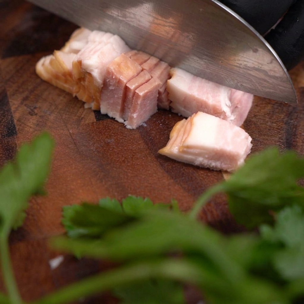 Un couteau bien aiguisé coupant une tranche de poitrine de porc crue pour créer de petits cubes uniformes sur une planche en bois foncé.