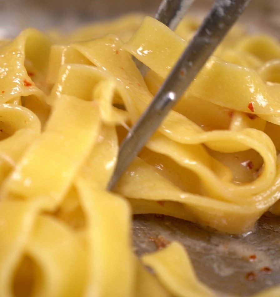 Des tagliatelles remuées avec des pinces en métal dans une poêle contenant des lardons et de leau de cuisson frémissante.