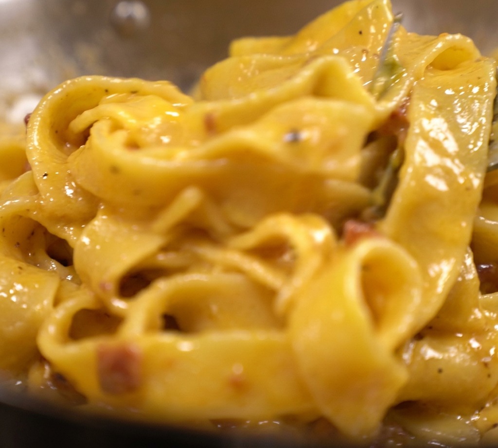 Des tagliatelles enrobées dune sauce aux œufs et au fromage crémeuse, épaisse et dun jaune éclatant, avec des lardons visibles dans la poêle.
