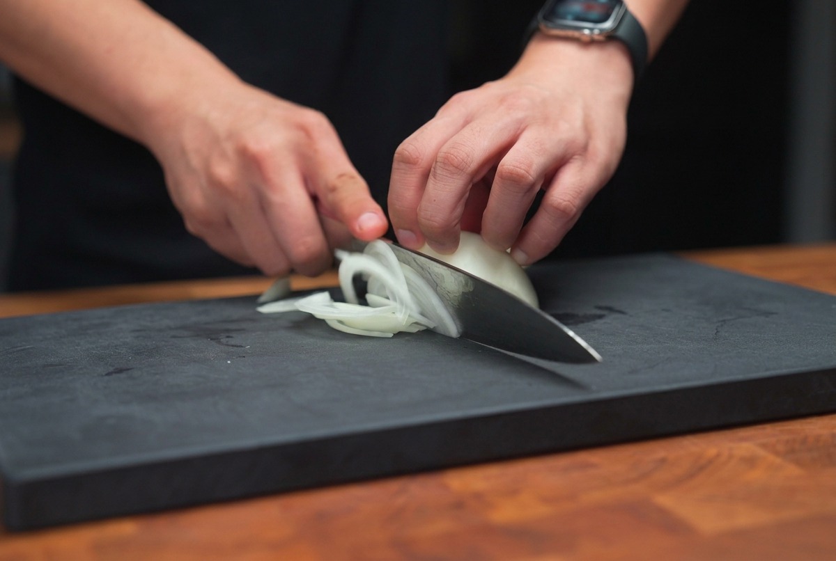 Mains tenant un couteau de chef, éminçant un oignon blanc en fines lamelles sur une planche à découper sombre.