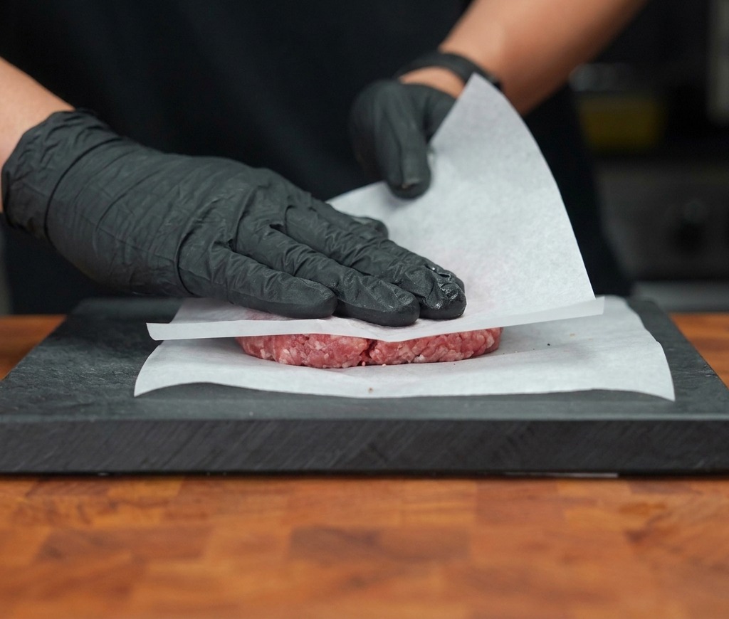 Mains gantées pressant fermement un steak haché cru entre deux feuilles de papier sulfurisé blanc sur une planche à découper.