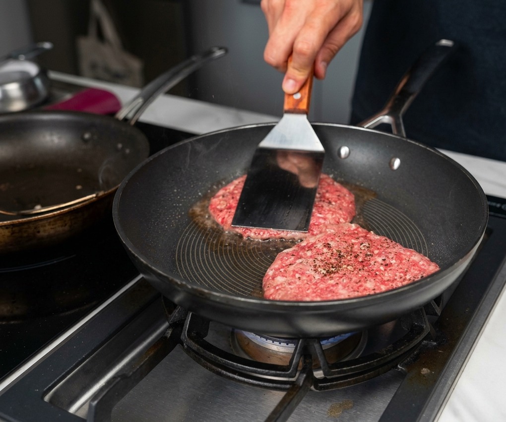 Deux steaks de bœuf saisis dans une poêle chaude, avec une spatule en métal pressant lun deux contre la surface de cuisson.