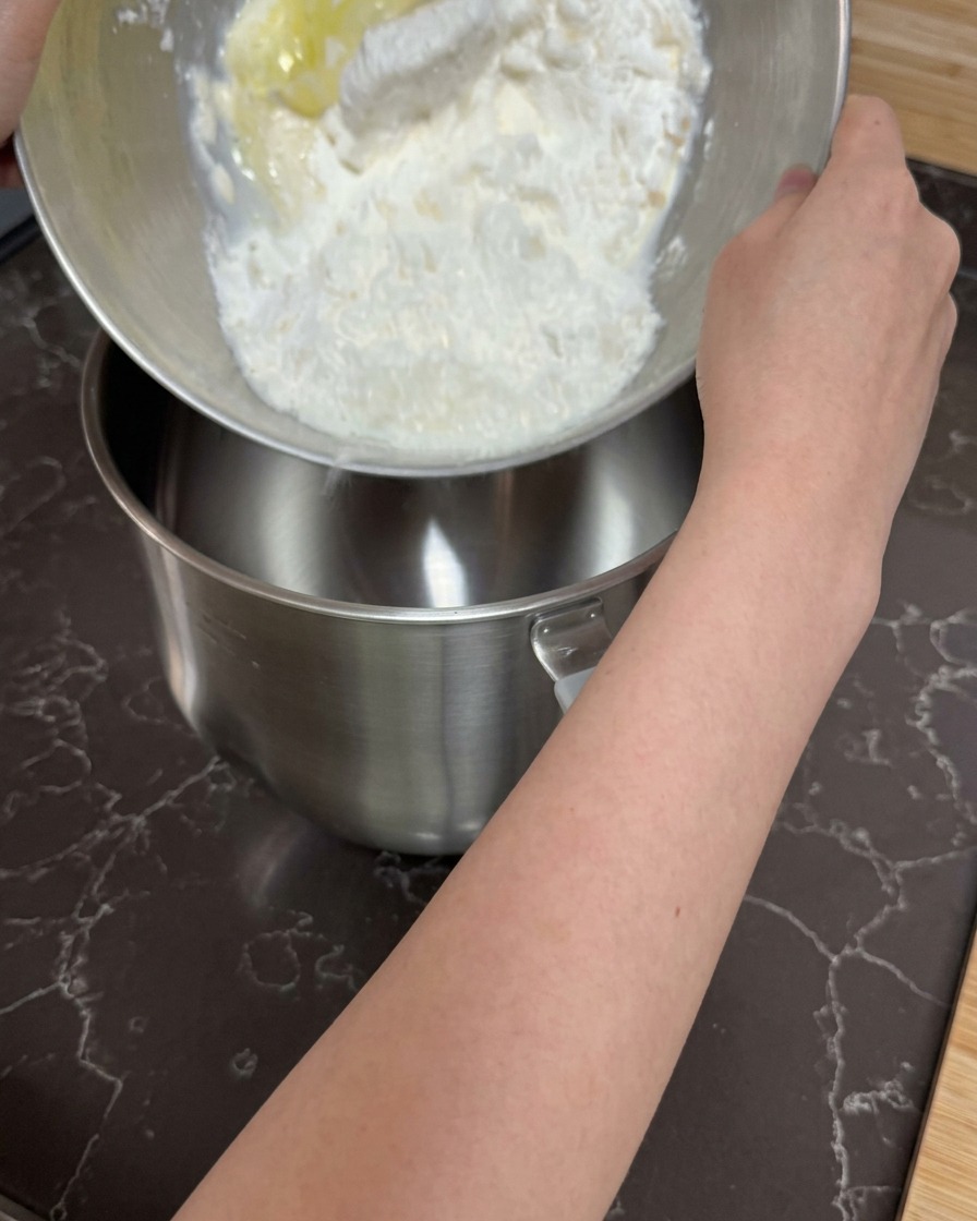 Des mains versant un mélange grossier de farine et dingrédients humides dans le bol vide dun robot pâtissier.